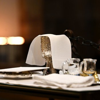 Kelch mit einem Tuch abgedeckt, daneben zwei kleine Glaskännchen mit Wasser und Weißwein