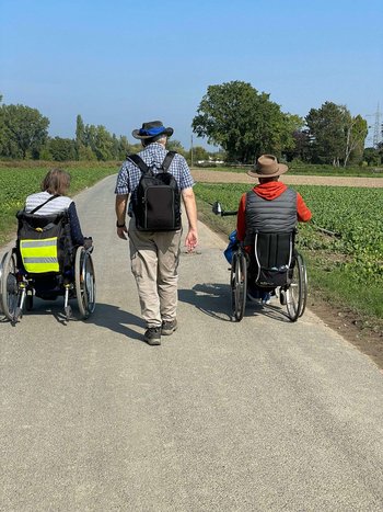 2 Rollstuhlfahrer und ein Fußgänger sind auf einem Weg unterwegs.