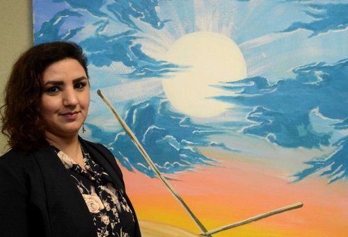 Eine Frau steht vor einem Gemälde. Es zeigt im oberen Teil einen weiß-blauen Himmel mit Streifen, die in eine hellgelbe Sonne hineinreichen. Der untere Teil des Bildes ist gelb-orange gefärbt und man sieht zwei Stäbe, die in die Fläche hineinragen.