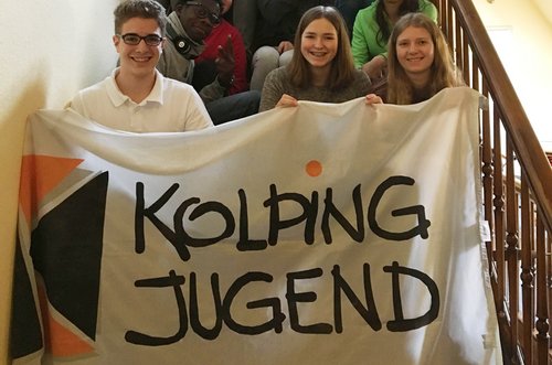 Drei junge Menschen halten Banner mit Logo der Kolpingjugend hoch.