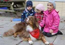 Hund mit rot-weißem Halstuch mit Logo der Malteser liegt auf einem Weg, dahinter sitzen drei kleine Kinder.