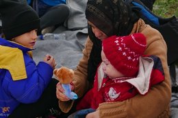 Eine Frau mit Kopftuch sitzt mit kleinem Kind im Arm auf dem Boden und zeigt dem Kind ein Kuscheltier. Daneben sitzt ein etwas älteres Kind und guckt zu.