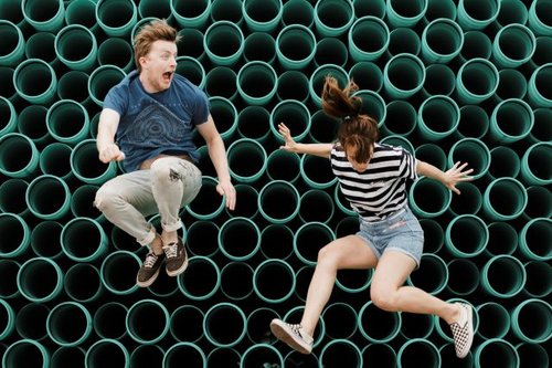 Ein junger Mann und eine junge Frau springen vor einer Wand mit lauter Röhren hoch.