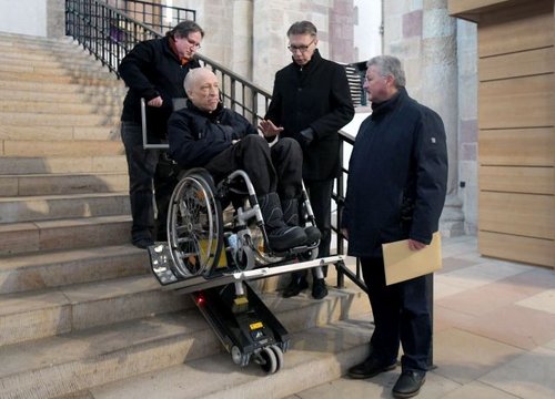 Mann im Rollstuhl wird mit einer Treppenraupe die Treppe zum Querhaus im Speyerer Dom hochtransportiert.