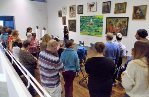 Eine Gruppe von Menschen steht in einem großen Raum vor einer Wand mit Gemälden. Vorne steht eine Frau, die die Bilder erklärt.