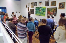 Eine Gruppe von Menschen steht in einem großen Raum vor einer Wand mit Gemälden. Vorne steht eine Frau, die die Bilder erklärt.