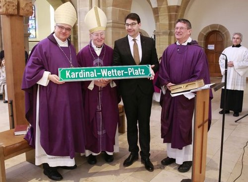 Vier Männer stehen in einer Kirche: Bischof Dr. Karl-Heinz Wiesemann, Kardinal Friedrich Wetter, OB Thomas Hirsch. Dekan Axel Brecht. Sie halten ein Schild mit der Aufschrift "kardinal-Wetter-Platz".